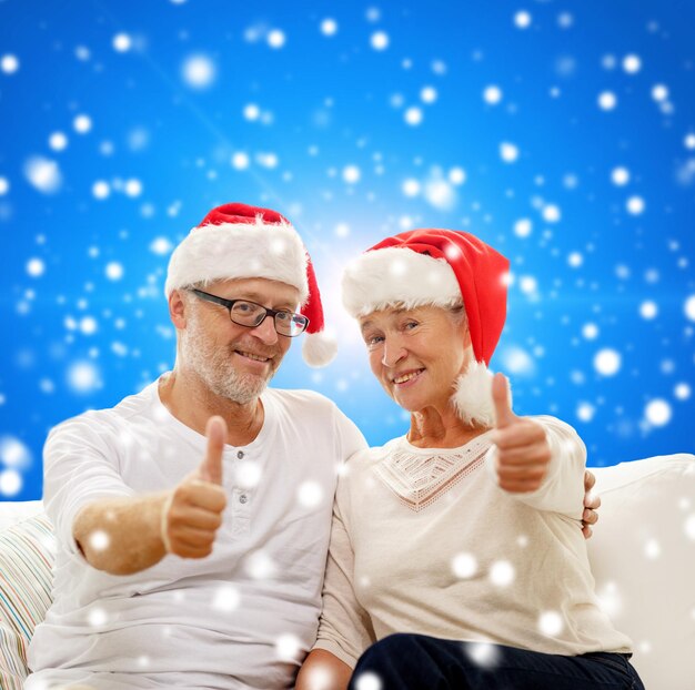 family, holidays, christmas, age and people concept - happy senior couple in santa helper hats sitting on sofa over blue snowy background