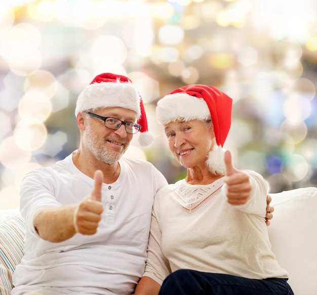 family, holidays, christmas, age and people concept - happy senior couple in santa helper hats sitting on sofa over lights background and showing thumbs up gesture