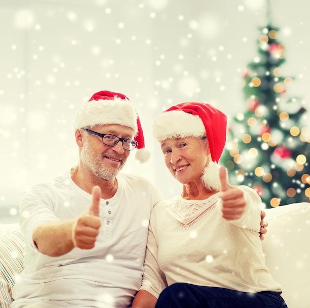 family, holidays, christmas, age and people concept - happy senior couple in santa helper hats sitting on sofa over living room and christmas tree background