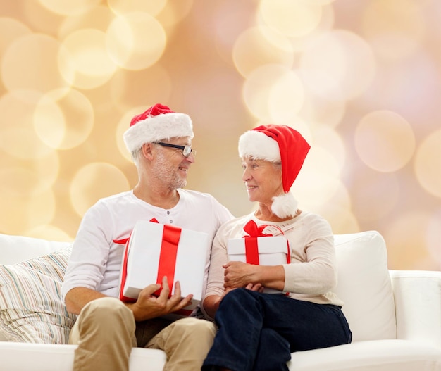 family, holidays, christmas, age and people concept - happy senior couple in santa helper hats with gift boxes over beige background