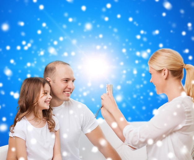 family, holidays, christmas technology and people concept - smiling mother, father and little girl with camera over blue snowy background