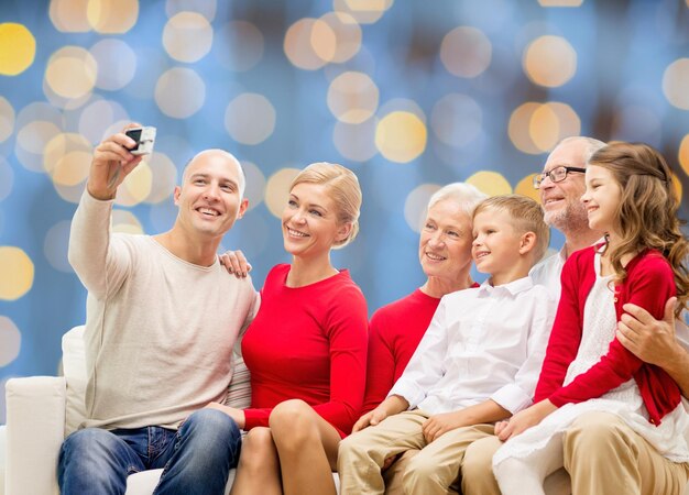 Photo family, holidays, generation, christmas and people concept - smiling family with camera taking selfie and sitting on couch over blue lights background