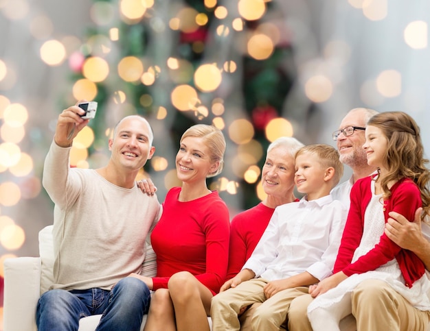 family, holidays, generation, christmas and people concept - smiling family with camera taking selfie and sitting on couch over tree lights background