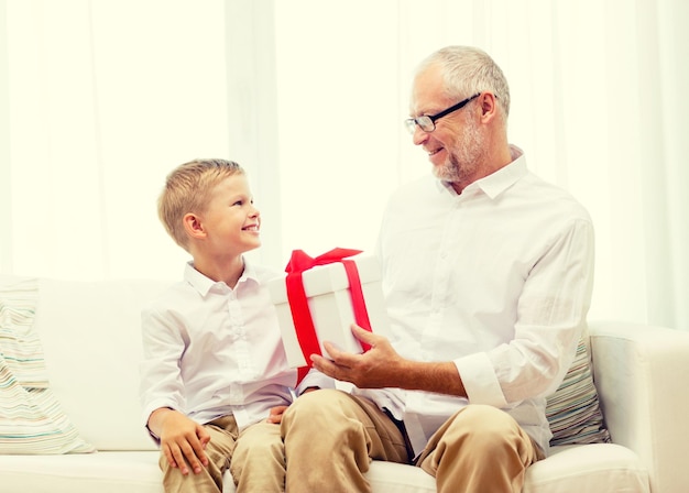 family, holidays, generation, christmas and people concept - smiling grandfather and grandson with gift box sitting on couch at home