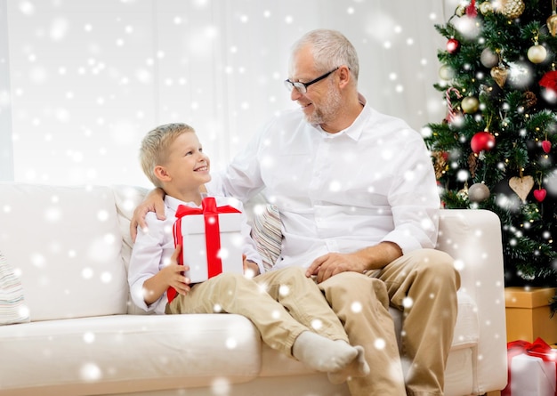 семья, праздники, поколение, рождество и концепция людей - улыбающиеся дедушка и внук с подарочной коробкой сидят дома на диване