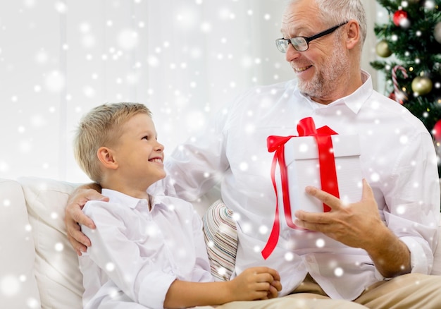 family, holidays, generation, christmas and people concept - smiling grandfather and grandson with gift box sitting on couch at home