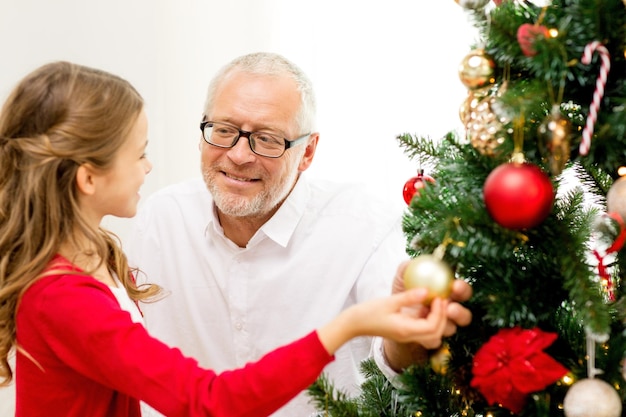 family, holidays, generation and people concept - smiling girl with grandfather decorating christmas tree at home