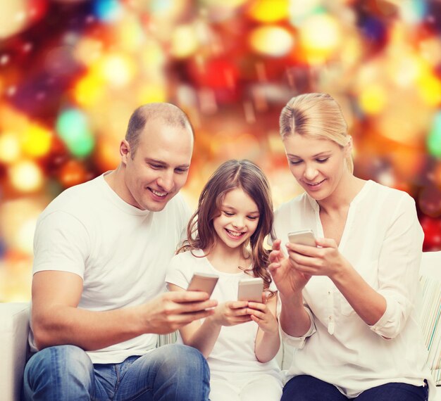 Photo family, holidays, technology and people concept - smiling mother, father and little girl with smartphones over red lights background