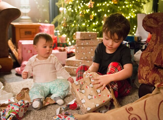 クリスマスの日に家にいる家族2人の子供が木のそばのプレゼントの間に座っています