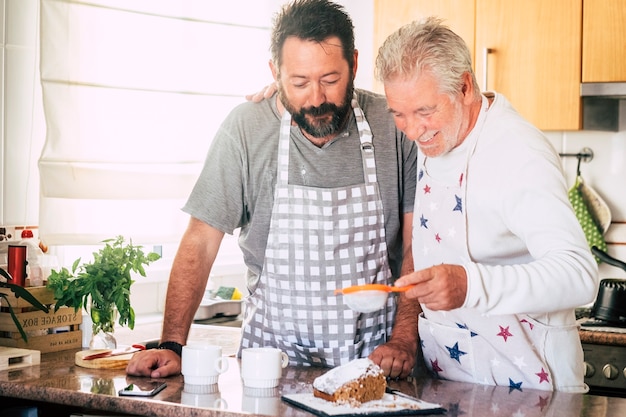 Famiglia a casa al lavoro in cucina con padre maturo anziano e figlio adulto che preparano una torta insieme come amici - diversità e generazioni miste in casa