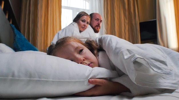 A family in the hotel room a girl sleeping in bed her mom and dad looking at her