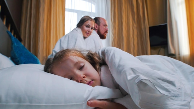 A family in the hotel room a little girl sleeping in bed her mom and dad looking at her