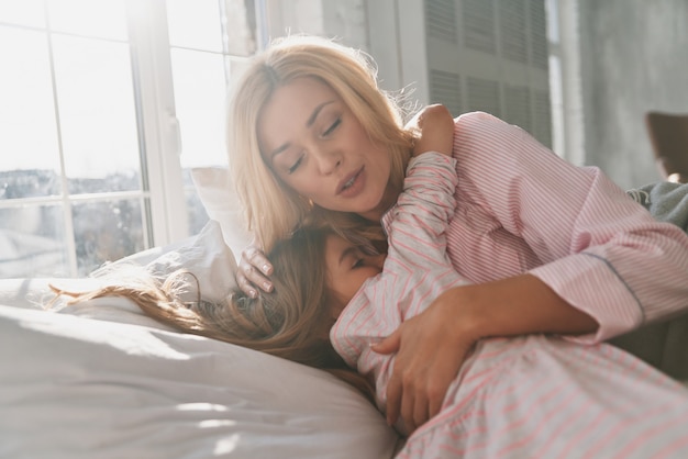 Family is the biggest treasure. Young beautiful mother with her cute little daughter embracing and smiling while lying on the bed at home