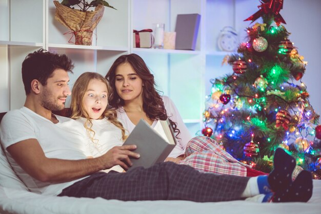 The family lay on the bed with a book near the christmas tree. evening time