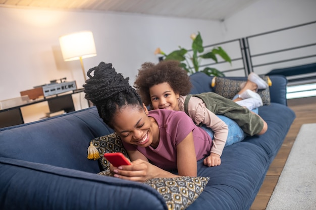 Family leisure. Laughing darkskinned young woman with smart phone and little curly daughter lying on sofa at home cheerful spending leisure time