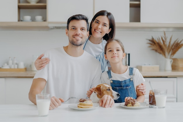 Family leisure pastime eating concept Father mother and daughter jack russell terrier dog pose all together at camera against kitchen interior get pleasure from eating pancakes drink fresh milk