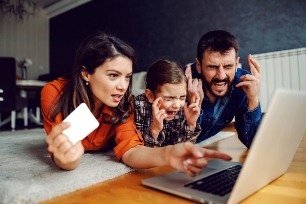 Family lying on the floor and using laptop for online shopping. Mom holding credit card and pointing at laptop while dad and daughter holding fingers crossed for luck.