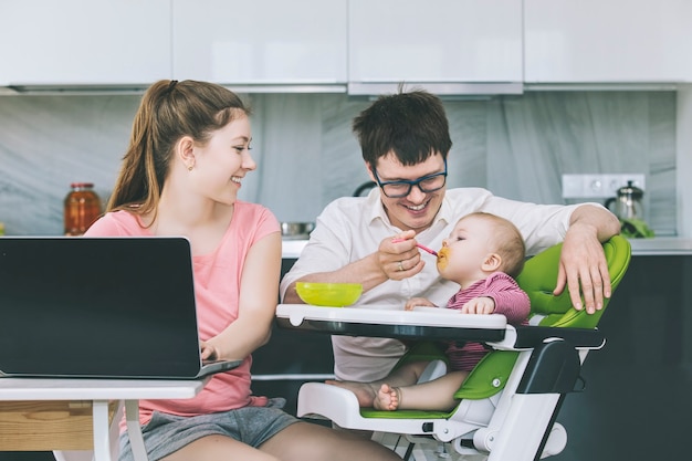 Family mom and dad feeding baby in the kitchen happy together at home smiling in the kitchen