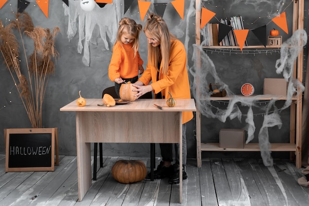 Foto mamma e figlia di famiglia preparano la zucca per halloween a casa in una stanza decorata ragazze bionde stanno smistando una zucca con un cucchiaio buona famiglia di halloween