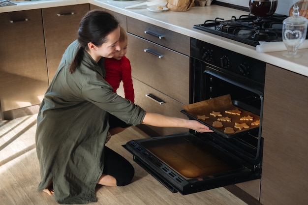 Family mom with toddler baby girl cooking gingerbread and puts cookies in oven christmas homemade