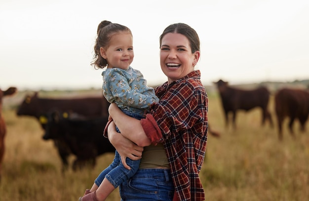 Family mother and baby on a farm with cows in the background eating grass sustainability and agriculture Happy organic dairy farmer mom with her girl and cattle herd outside in sustainable nature