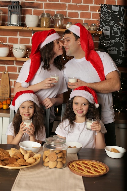 Foto madre di famiglia, padre e 2 figlie in cucina con cappelli di babbo natale e latte in mano