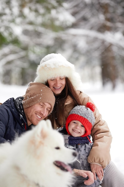 Family of mother father and son having fun in snowy winter wood with cheerfull pet dog