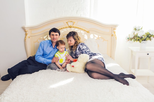 家族、親子関係、子供のコンセプト-幸せな母、父と息子が自宅の寝室のベッドでテディベアと一緒に遊んでいます。