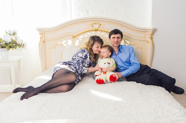 Family, parenthood and children concept - Happy mother, father and son playing together with teddy bear on bed in bedroom at home.