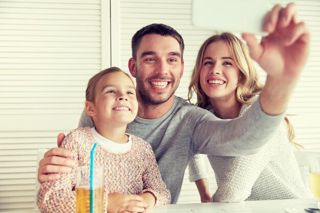 가족, 부모, 기술, 사람 개념 - 행복한 어머니, 아버지, 어린 소녀가 저녁 식사를 하고 레스토랑에서 스마트폰으로 셀카를 찍는다