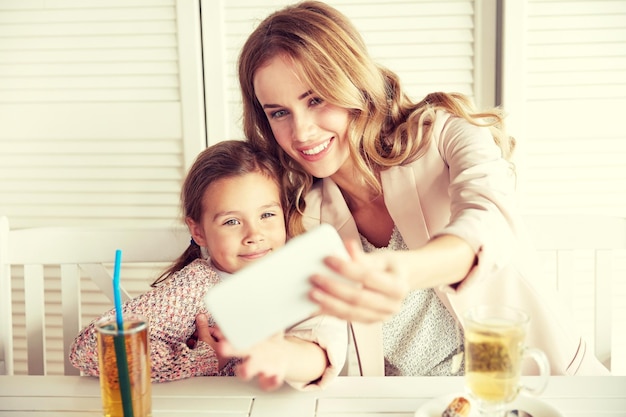 family, parenthood, technology and people concept - happy mother and little girl having dinner and taking selfie by smartphone at restaurant