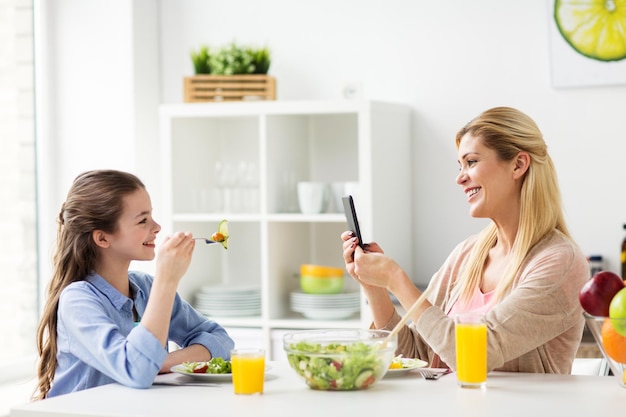 家族と人々のコンセプト – スマートフォンで夕食をとり、自宅のキッチンで娘の写真を撮る幸せな母親