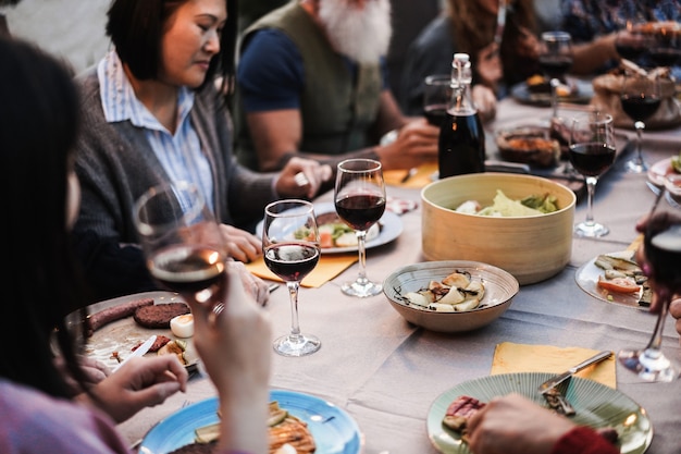 Family people having fun at barbecue dinner - multiracial\
friends eating at bbq meal outdoor - food, friendship, gathering\
and summer lifestyle concept - focus on center wine glass