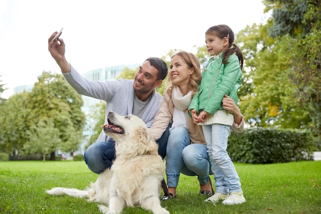 家族、ペット、動物、技術、人のコンセプト – ラブラドル・レトリーバー犬が公園でスマートフォンで自撮りをする幸せな家族