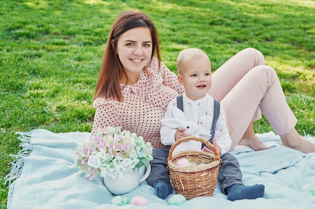 Family photo session of mom and baby son for Easter in the park, next to them is a basket with eggs and an Easter rabbit