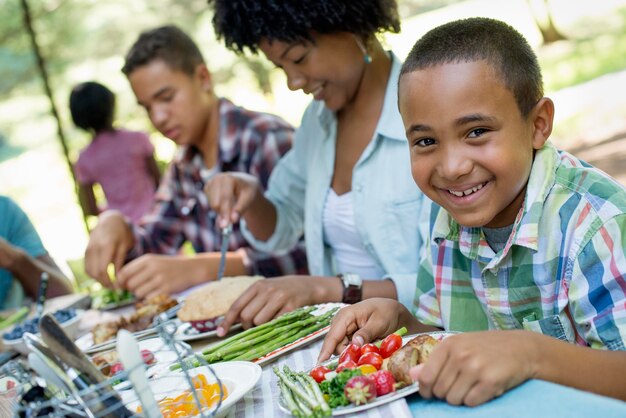 A family picnic meal in the shade of tall trees Parents and children helping themselves to fresh fru