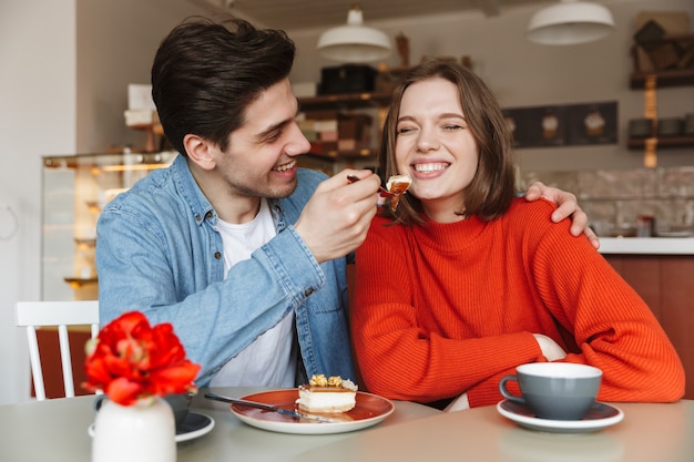 Family portrait of happy couple eating sweets, while man feeding woman with tasty cake in cafe