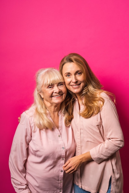 Family portrait, middle aged woman and senior mother embracing on pink wall