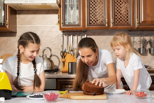 Family portrait of mom and daughters are preparing birthday cake together, mom is cutting sponge cake using knife in kitchen.