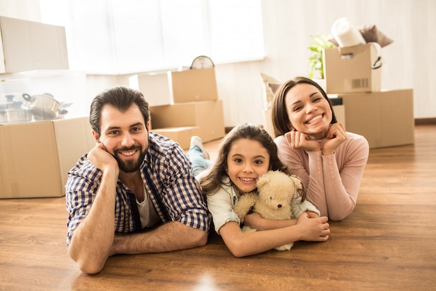 Family portrait of three people lying on the floor. Man, woman and their daughter looks cheerful and happy. There are lots of boxes behind them.