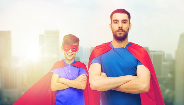 family, power and people concept - man and boy wearing mask and red superhero cape over city skyscrapers background