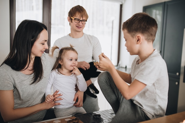 La famiglia prepara i biscotti di pan di zenzero in cucina. papà, mamma, figlio, figlia, fratello e sorella. impastare, tagliabiscotti, capodanno, natale, cena festiva. abbigliamento per la casa grigio.