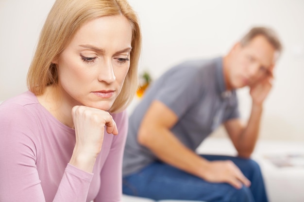 Family problems. Side view of depressed mature woman holding hand on chin while sad man sitting on background