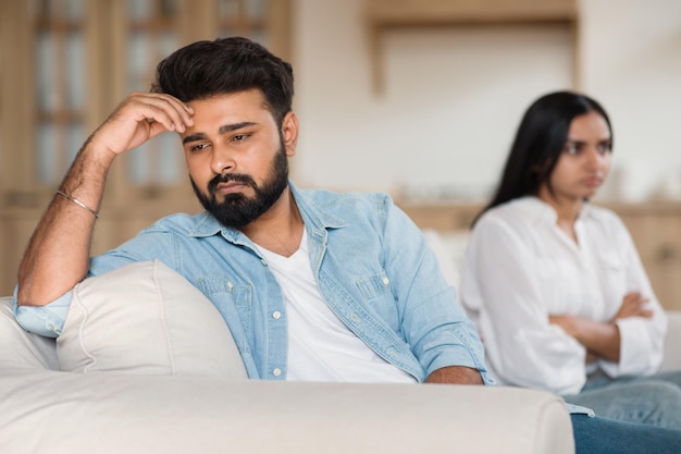 Photo family problems unhappy indian man sitting separate from discontented wife on sofa at home couple