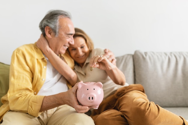 Family savings happy senior couple putting coin in piggybank while sitting together on sofa at home