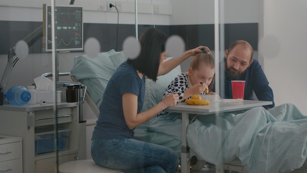 Family sitting with sick little daughter while eating healthy
nutrition food during recovery examination in hospital ward. child
patient wearing oxygen nasal tube resting in bed having meal
lunch