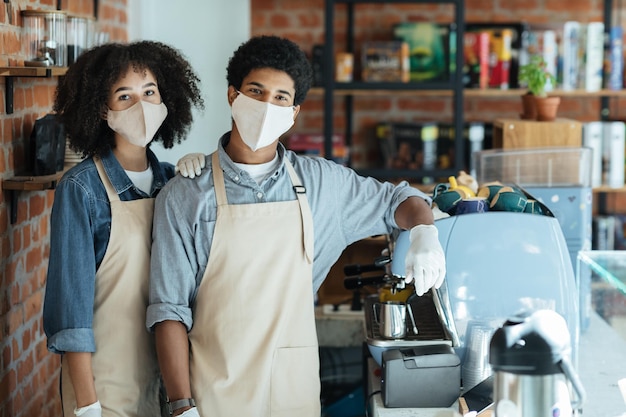 Family small business and work during COVID-19 epidemic. Millennial african american couple in apron, protective mask and gloves look at camera in interior of modern coffee shop with equipment