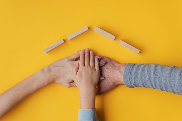 Family stacking their hand on yellow wall with roof made of wooden blocks above their hands in a conceptual image of home ownership and safety