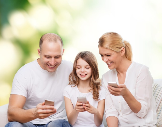 семья, лето, технологии и люди - улыбающиеся мать, отец и маленькая девочка со смартфонами на зеленом фоне