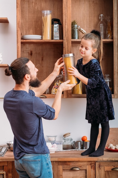 Family teamwork. Father cooking in kitchen with cute little assistant. Young daughter helping to get jar with spaghetti from shelf.